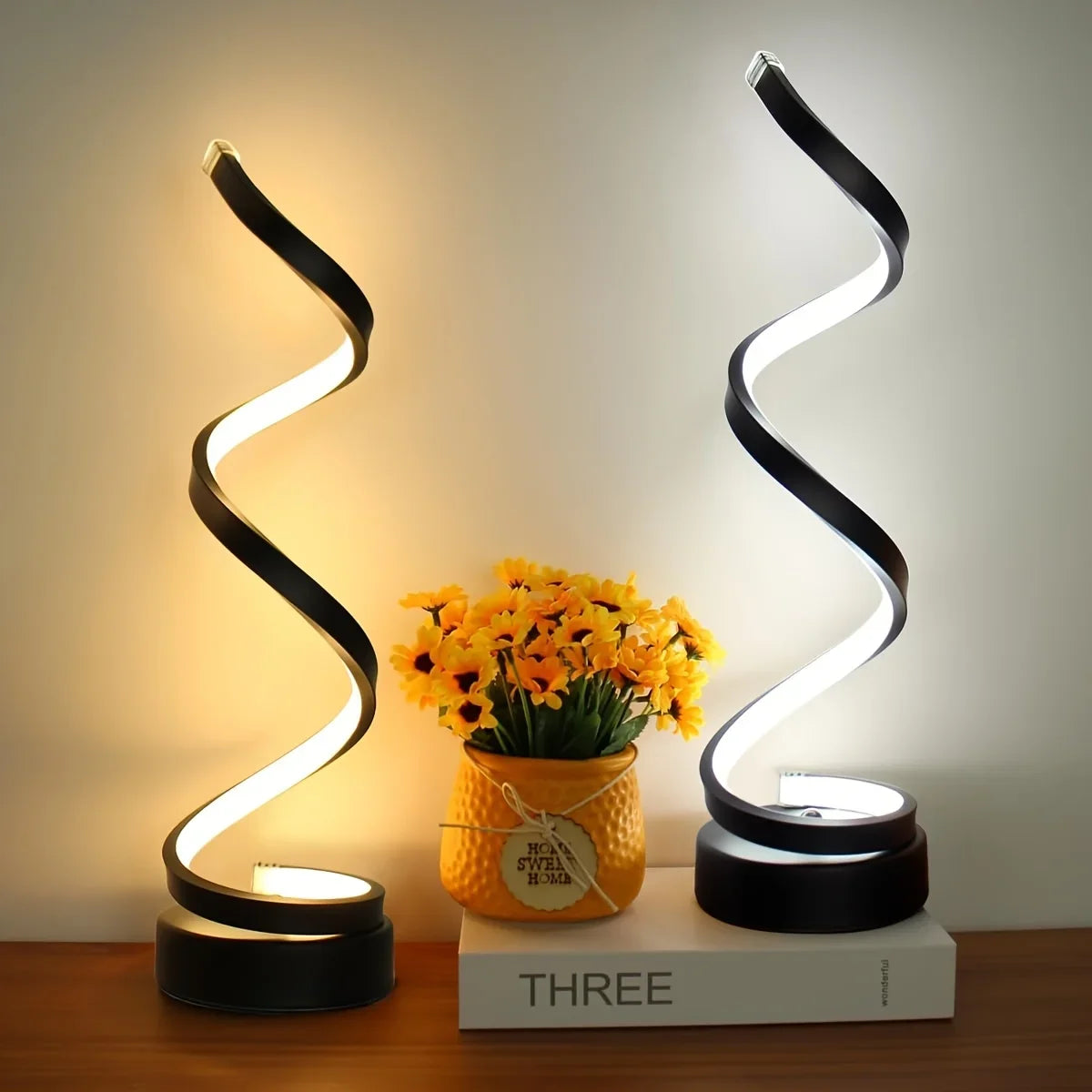 Pair of modern spiral LED lamps in matte black, emitting warm and cool light tones, placed symmetrically on a tabletop beside yellow flowers, creating a balanced ambient aesthetic, ThuisGemak.