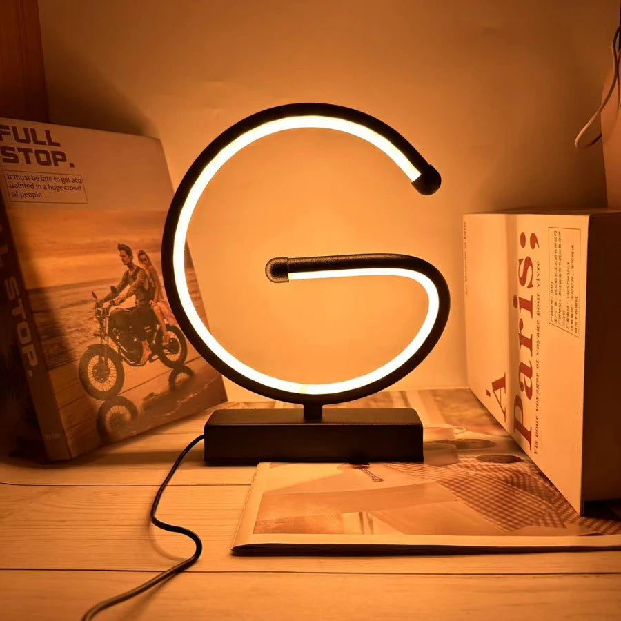 Modern LED table lamp shaped like the letter G, in matte black with a warm amber glow, placed on a wooden surface beside books and decor, creating a cozy ambient light, ThuisGemak.